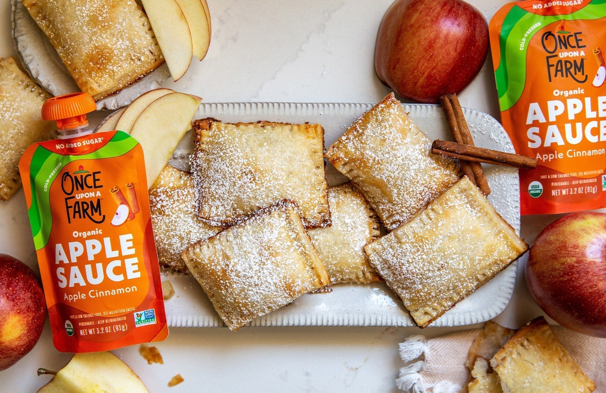 apple turnovers on a plate, surrounded by apples, cinnamon sticks, and Once Upon a farm applesauce pouches