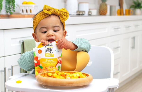 baby in highchair, holding a package of Once Upon a Farm frozen baby food, with the cooked meal on her plate