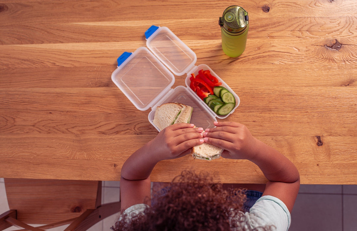 top-down view of a child, sitting at a table and holding a sandwich over a lunchbox