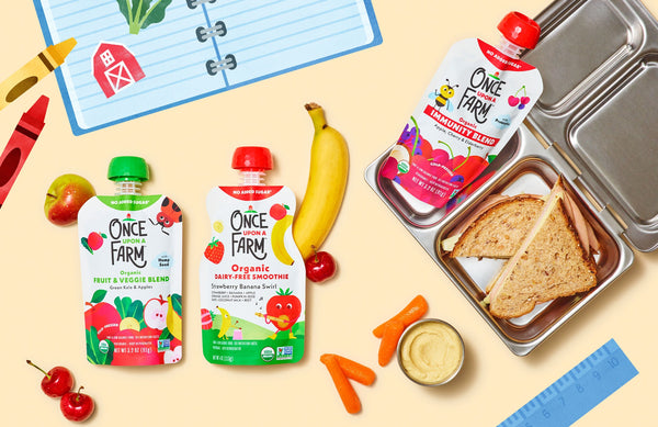 metal lunchbox with a sandwich and Once Upon a Farm pouch inside. Next to it are fresh fruits and veggies and two more Once Upon a Farm pouches